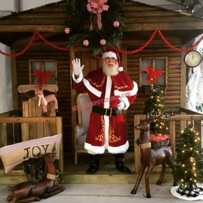 Santa Claus impersonator waving in front of a log cabin Christmas set with reindeer and a JOY sign